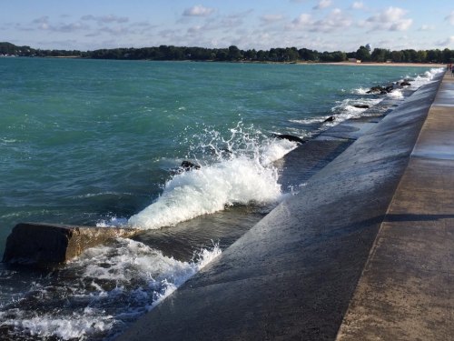 Waves crash against a rocky shoreline under a blue sky with scattered clouds, near a calm body of water.