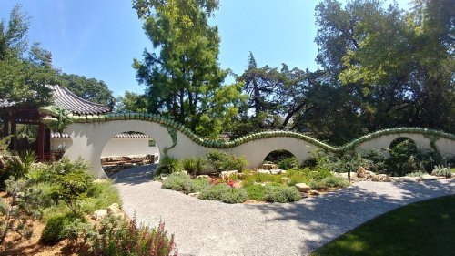 A serene garden with a curved dragon wall, lush greenery, and a clear blue sky in the background.