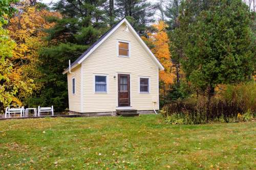 A small yellow house surrounded by trees with autumn foliage, featuring a front porch and a grassy yard.