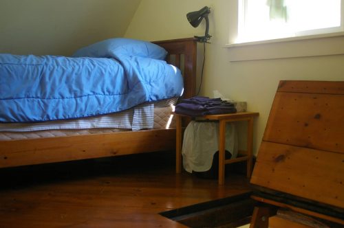A cozy bedroom with a blue comforter on a bed, a wooden nightstand, and a window letting in natural light.