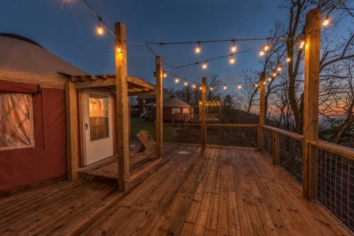 Cozy wooden deck with string lights, leading to a yurt at dusk, surrounded by trees and a scenic view.