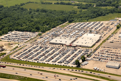 Aerial view of a large parking lot filled with cars next to a commercial building and green fields.