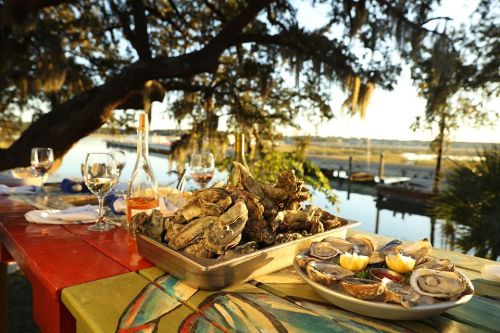 A scenic outdoor dining setup with oysters, wine, and a view of the water at sunset.