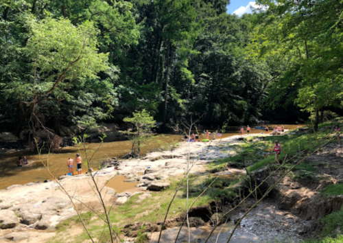A scenic riverbank with people enjoying the water, surrounded by lush greenery and rocky terrain.