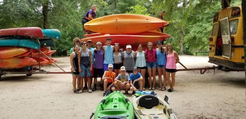 A group of young people poses in front of stacked kayaks and a yellow bus at a outdoor adventure site.