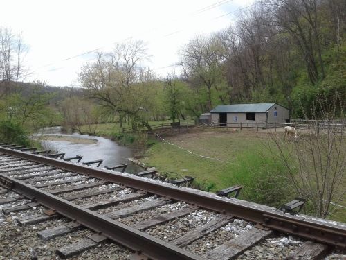 A peaceful scene featuring a river, a barn, and a horse near a railway track surrounded by trees.