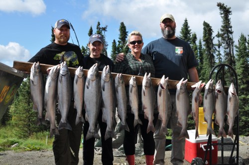 Four people stand together, smiling, holding a line of freshly caught fish in a forested outdoor setting.