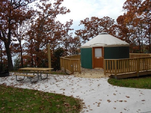 A yurt with a wooden deck surrounded by autumn trees and fallen leaves, featuring a picnic table nearby.