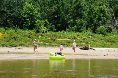 Three people play volleyball on a sandy beach near a river, with a bright yellow inflatable raft nearby.