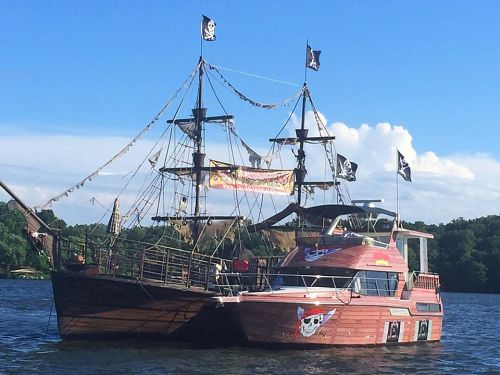 A pirate-themed boat with flags sailing on a lake under a clear blue sky.