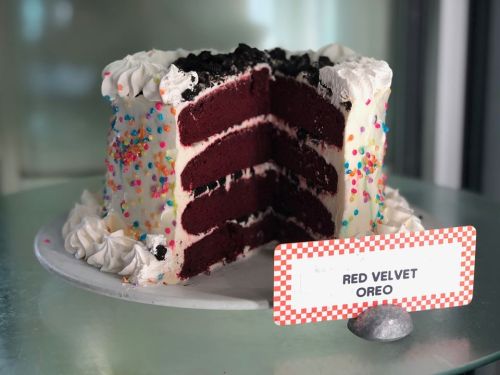 A red velvet cake with layers, topped with cream and colorful sprinkles, displayed on a stand with a label.