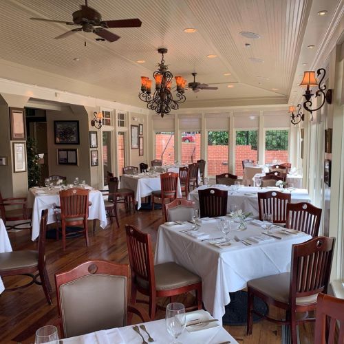 Elegant restaurant interior with white tablecloths, wooden chairs, and warm lighting, featuring large windows and a chandelier.
