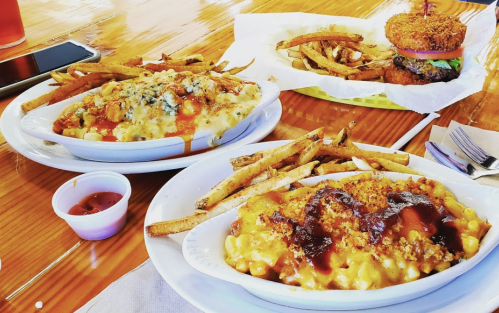 Two plates of loaded mac and cheese with fries, and a burger with onion rings in the background on a wooden table.