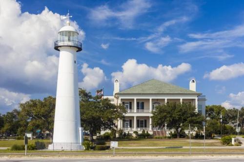 A white lighthouse beside a historic building under a blue sky with fluffy clouds. Green trees line the foreground.