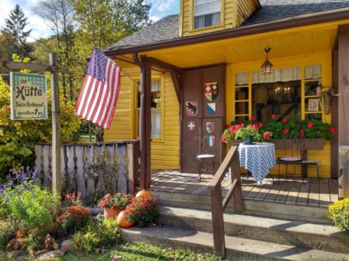A charming yellow house with a porch, American flag, and flower pots, featuring a sign for a Swiss restaurant.