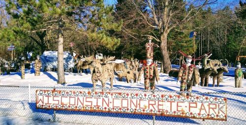 A snowy park featuring whimsical concrete sculptures and a sign reading "Wisconsin Concrete Park." Trees in the background.