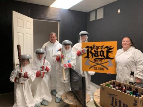 A group of six people in protective suits and helmets, holding bats and a sign that says "Age of Rage," in a room.