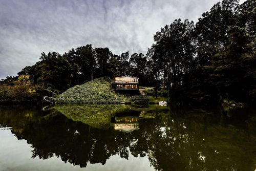 A modern house on a hill by a calm lake, surrounded by trees, reflecting in the water under a cloudy sky.