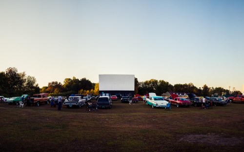 A vintage drive-in theater scene with classic cars parked on grass, people mingling, and a large screen in the background.