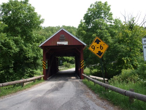 A red covered bridge surrounded by greenery, with a height clearance sign and a gravel road leading through it.