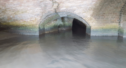 A dark, arched brick tunnel partially submerged in water, with reflections on the surface.