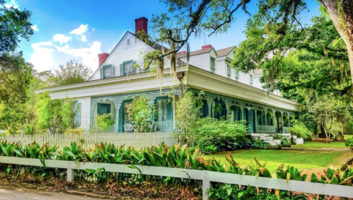 A charming two-story house with a wraparound porch, surrounded by lush greenery and trees under a bright blue sky.