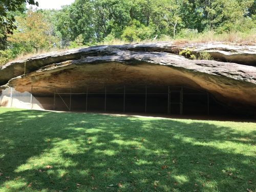 A large rock overhang with a fenced area beneath, surrounded by green grass and trees in the background.