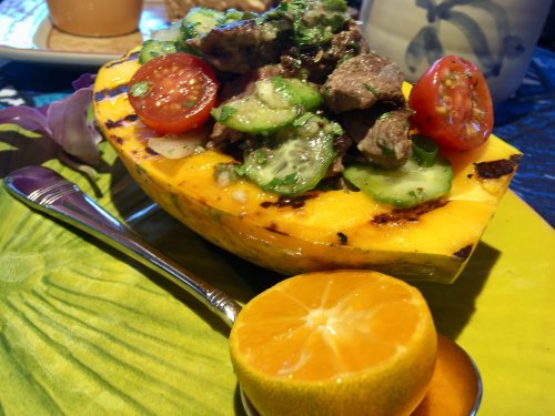 A grilled half papaya topped with beef, cucumbers, and cherry tomatoes, served with a slice of orange on a colorful plate.