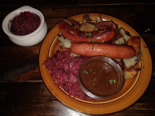 A plate with two sausages, sauerkraut, roasted potatoes, and a small bowl of red cabbage on a wooden table.