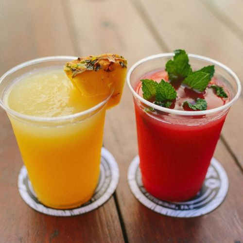 Two colorful drinks in clear cups: a yellow pineapple drink and a red beverage garnished with mint leaves.