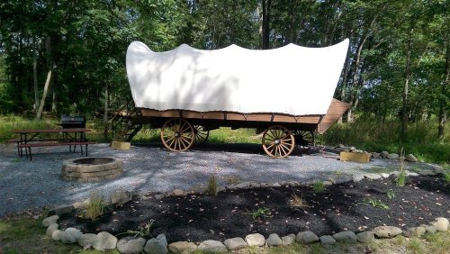 A covered wagon on wheels, set in a wooded area with a picnic table, fire pit, and landscaped stones around it.