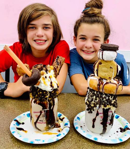 Two smiling kids sit at a table with oversized milkshakes topped with cookies, donuts, and whipped cream.
