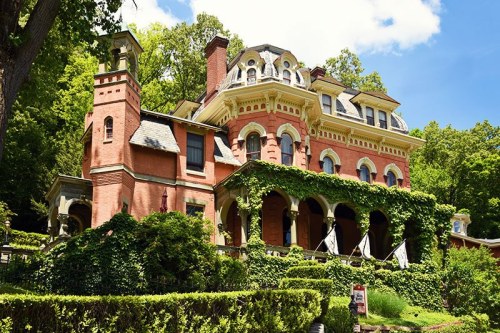 A large, ornate brick mansion surrounded by lush greenery and trees under a blue sky with fluffy clouds.
