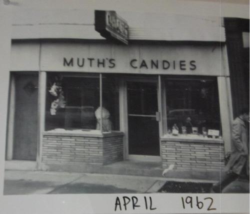 Black and white photo of Muth's Candies storefront, dated April 1962, featuring a brick facade and window displays.