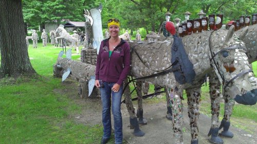 A woman stands beside a sculpture of a horse made from stones, with other artistic sculptures visible in the background.