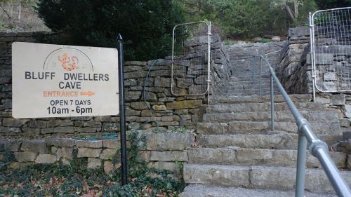 Sign for Bluff Dwellers Cave with stairs leading up, surrounded by stone walls and greenery. Open daily from 10am to 6pm.