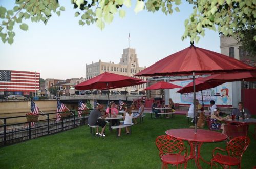 Outdoor seating area with red umbrellas, people dining, and American flags near a riverfront in a city setting.