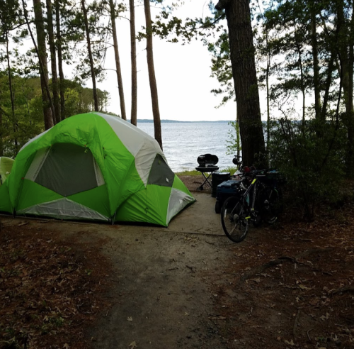 A green tent by a lake, surrounded by trees, with a bike and camping gear nearby on a dirt path.
