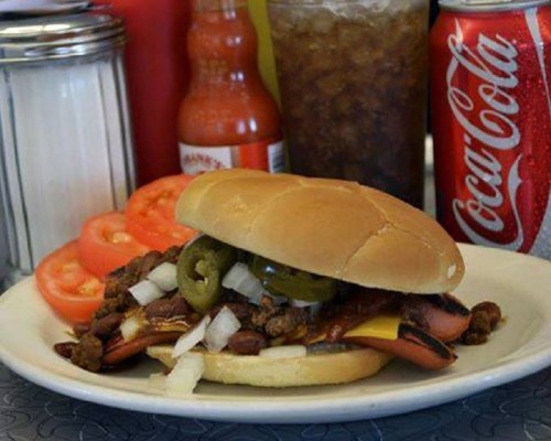 A hearty sandwich with ground meat, cheese, jalapeños, and onions, served with tomato slices and drinks in the background.
