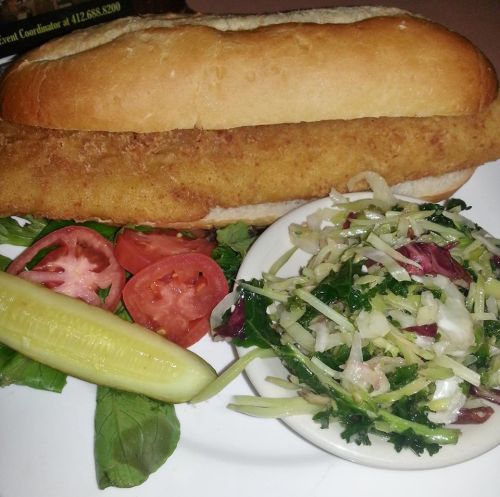 A crispy fish sandwich on a bun, served with sliced tomatoes, pickles, and a side salad.