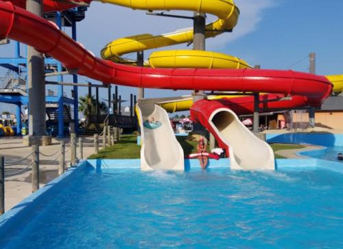 Two water slides in a pool area, with a child sliding down one and a lifeguard watching nearby under a clear blue sky.