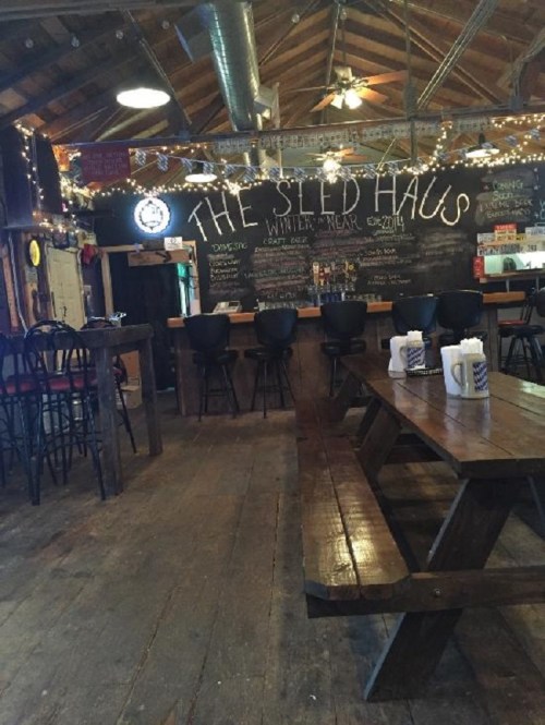 Interior of a rustic bar named "The Shed Haus," featuring wooden tables, a chalkboard menu, and string lights.