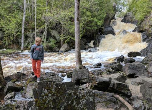 A child stands on rocks near a rushing waterfall surrounded by trees and greenery.