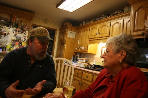 A man and woman engage in conversation at a kitchen table, with a glass of wine in front of the woman.