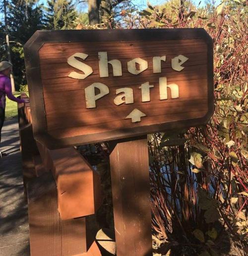 A wooden sign reading "Shore Path" with an arrow pointing up, surrounded by greenery and a walking path.