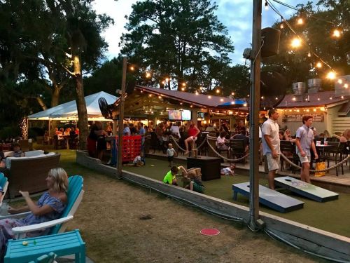 A lively outdoor venue with string lights, people socializing, and children playing on a grassy area.