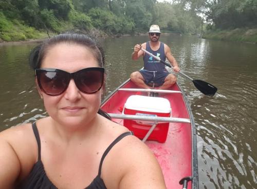 A woman in sunglasses takes a selfie while a man paddles a canoe on a calm river surrounded by greenery.