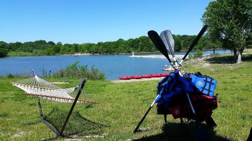 A serene lakeside scene with a hammock, kayak paddles, and a backpack, surrounded by lush greenery and clear blue skies.