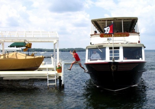 A person in a red shirt jumps from a dock onto a boat named "Walworth" on a sunny day.