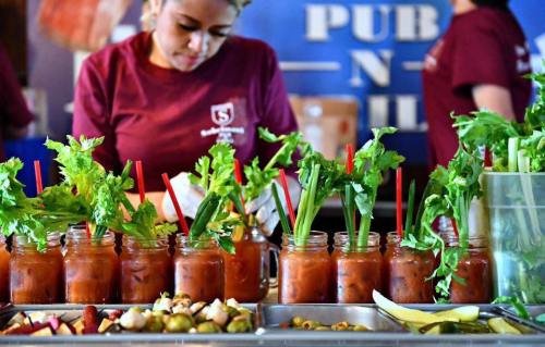 A woman prepares drinks with celery sticks in jars, surrounded by garnishes at a vibrant food event.